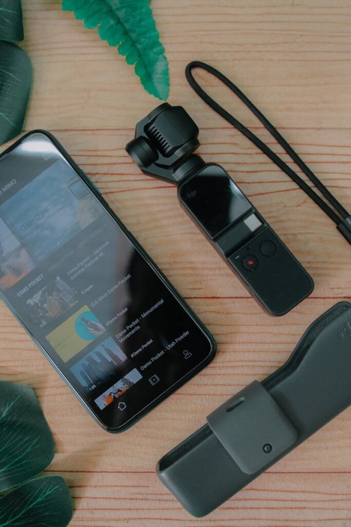A flat lay of a smartphone and camera with green leaves on a wooden table.